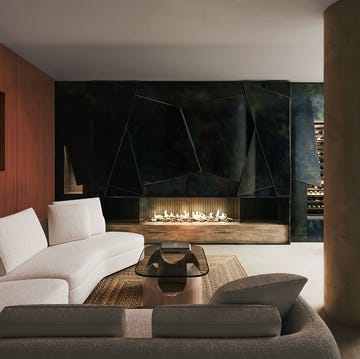 a living room with a wood paneled wall with a white 3d artwork, curved white sofa and a taupe colored loveseat, glass and stone cocktail table, supporting column, black zinc wall with fireplace, stone hearth
