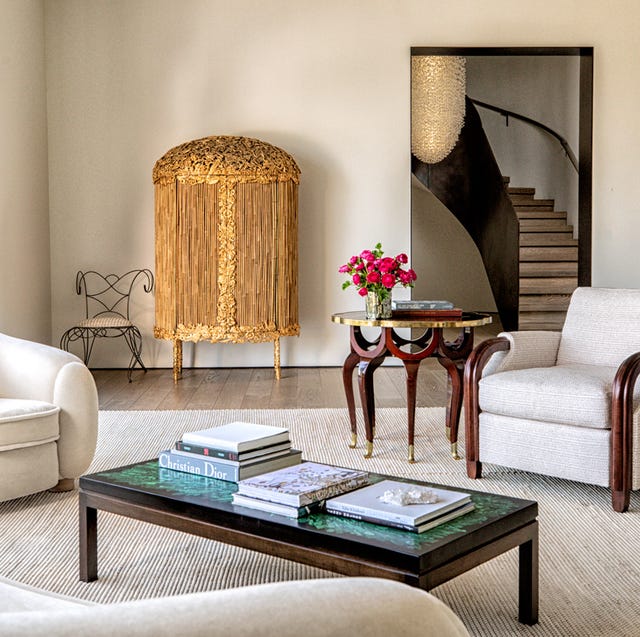 a living room with mismatched white armchairs and a swoopy curvy white sofa and rectangular cocktail table on a white striated rug and a gold encrusted rounded top console against the back wall where there is a doorway leading to a swoopy staircase upstairs