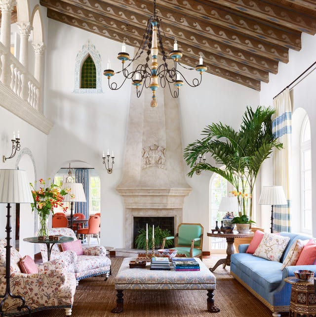 very dramatic high ceilinged living room with ornate wave designed beams up top and a tall fireplace hood and a wide upholstered cocktable table at center with a blue sofa on one side and two matching floral patterned armchairs to the left and a tall plume of palm tree at back and very long white and blue curtains next to arched windows
