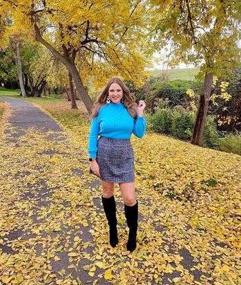 A reviewer stands on a leaf-covered path in a park, wearing a blue sweater, patterned skirt, and black knee-high boots