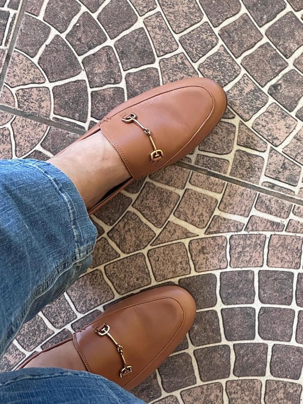 Person wearing leather loafers with metal buckle detail, standing on a stone-patterned floor, paired with casual jeans