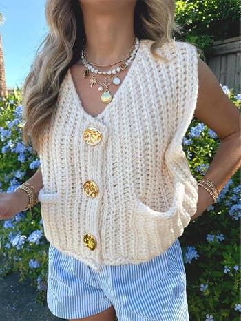 Model wearing a chunky knit vest with large buttons and striped shorts, layered necklaces, and bracelets, standing outdoors near flowers
