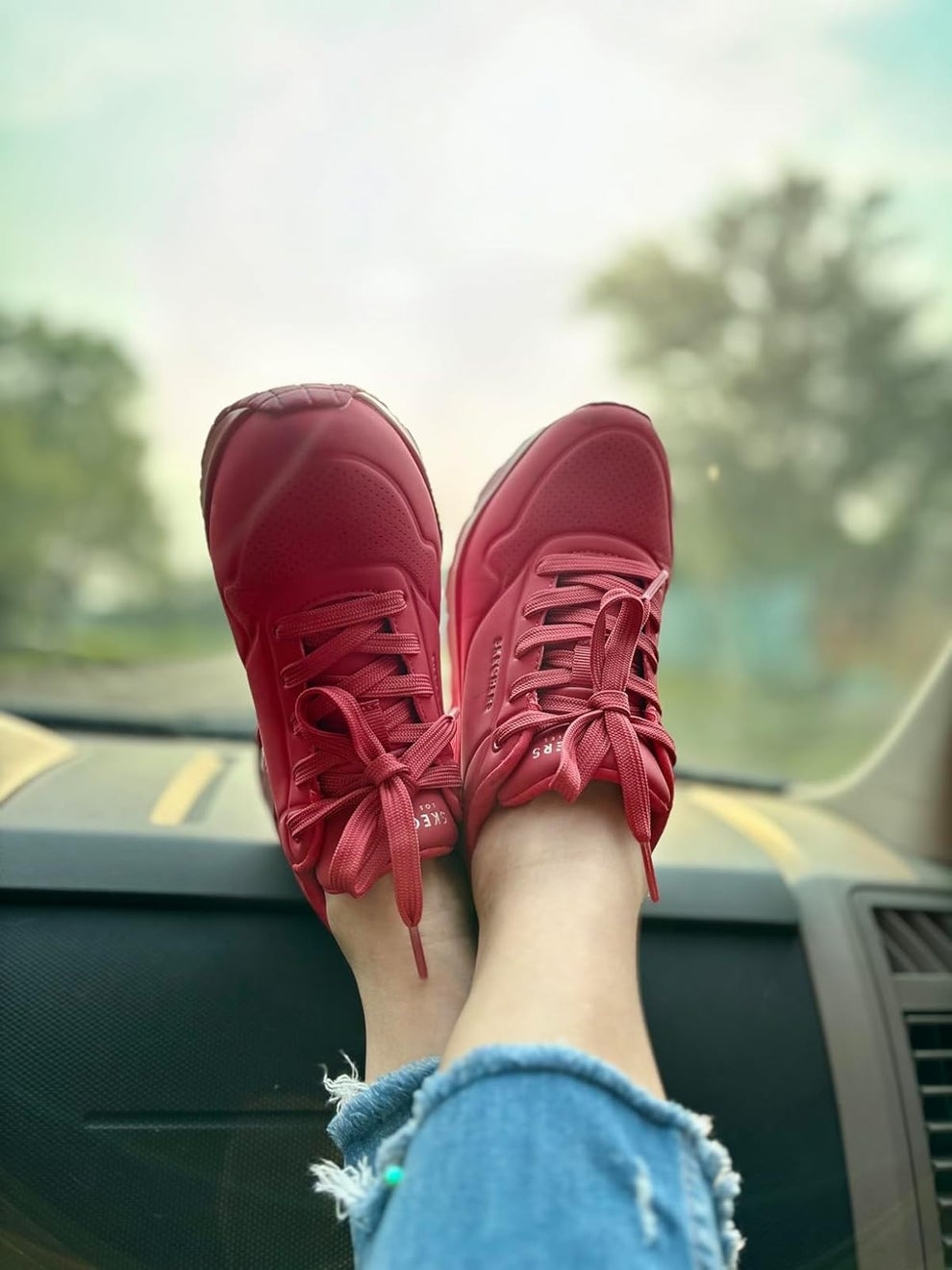 Feet wearing stylish sneakers rest on a car dashboard, with ripped jeans visible