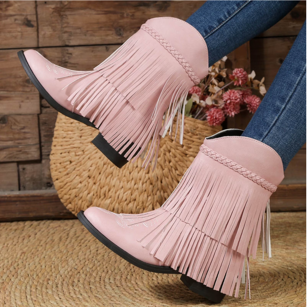 Fringed western-style boots with braided details, paired with denim jeans in a cozy room setting featuring rustic decor