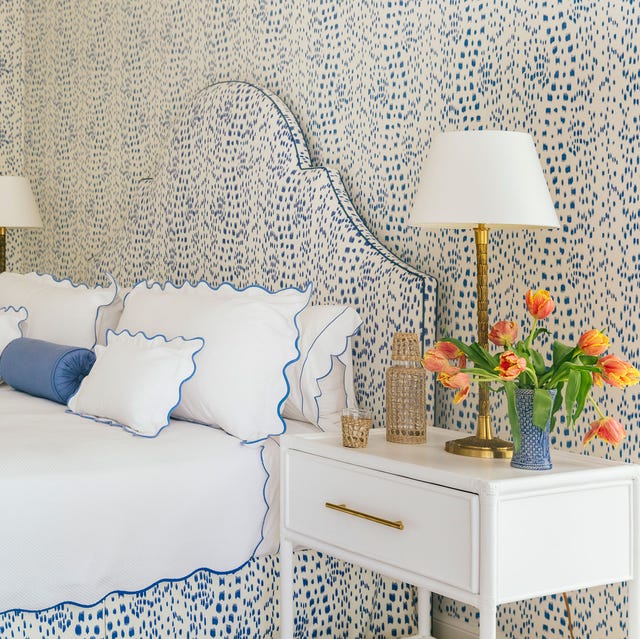 a bed with a white blue speckled bed headboard and matching wall and bed skirt and white linens edged in matching blue and a small side table with flowers and a lamp