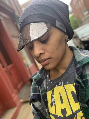Person wearing a patterned jacket, graphic tee, and a weather-resistant cap with a visor. They are looking down thoughtfully
