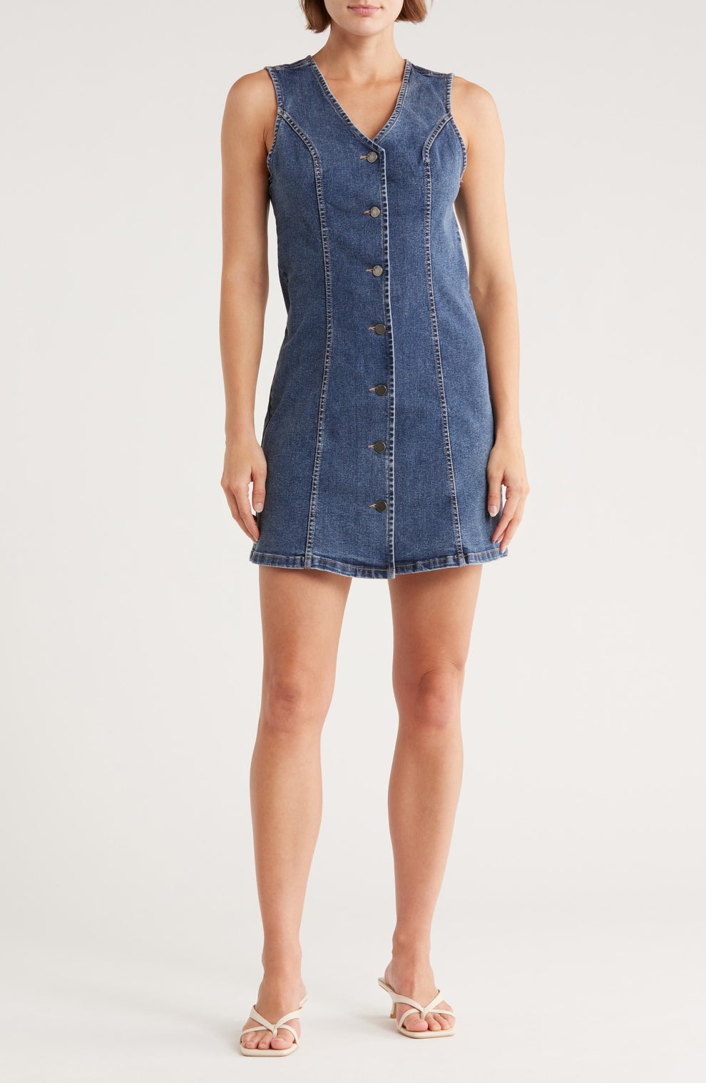 Person in sleeveless denim dress with front buttons and white sandals stands against a plain backdrop