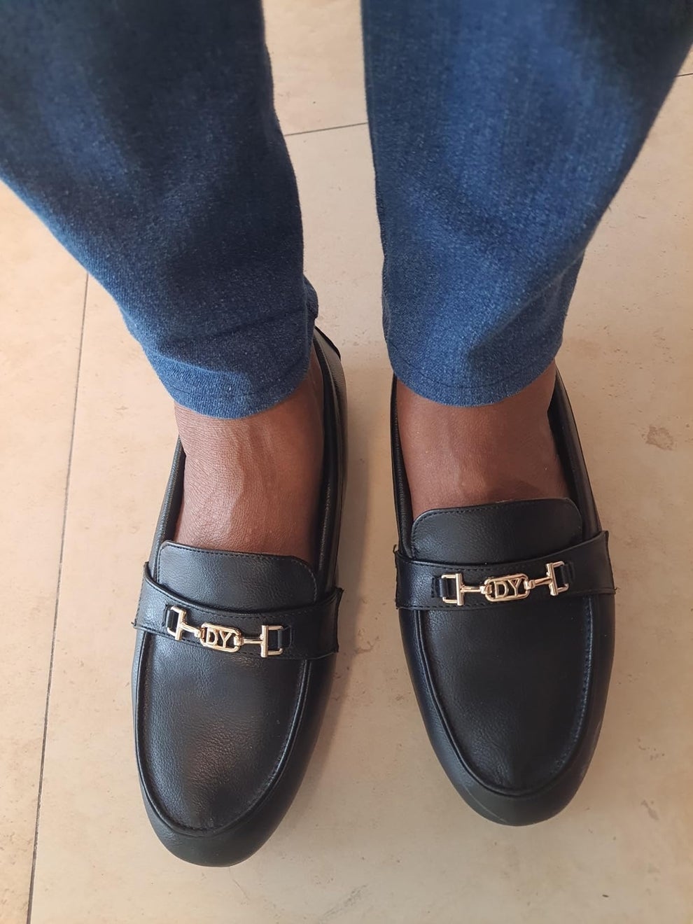 Person wearing stylish black loafers with metallic accent, paired with dark jeans, standing on a tiled floor