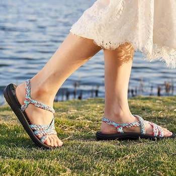 Person walking on grass near water, wearing braided sandals and a lace-trimmed dress