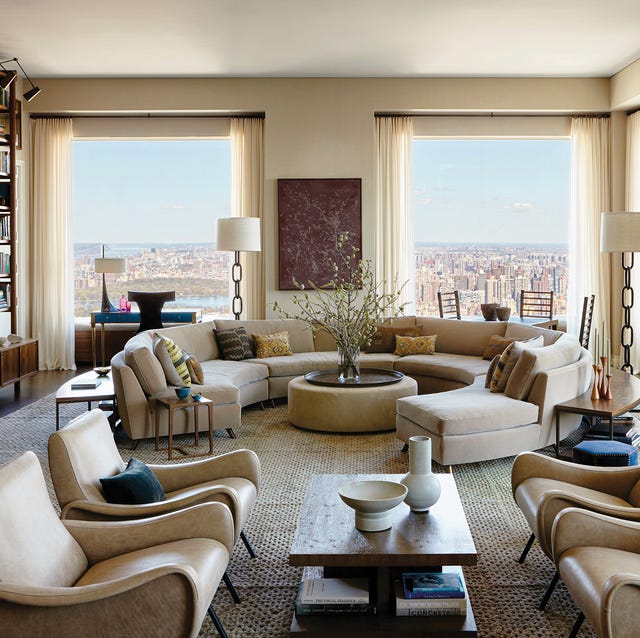 very off white and white living room with a nearly circular curvy sofa with round cocktail table ottoman at center and numerous chairs and tables and large high windows at back with a view of the city