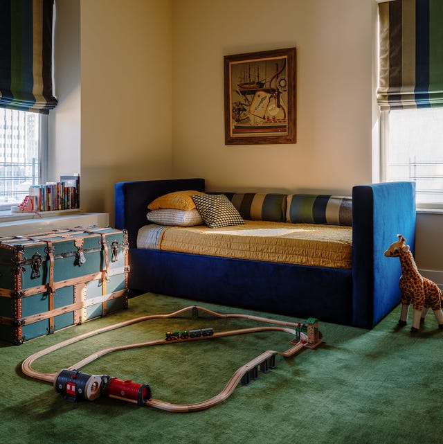 play room with blue velvet daybed with yellow cover and striped bolsters and an old blue strappy trunk to the left on a green carpet with a wooden train set and a small toy giraffe on the floor next to the daybed just hanging out