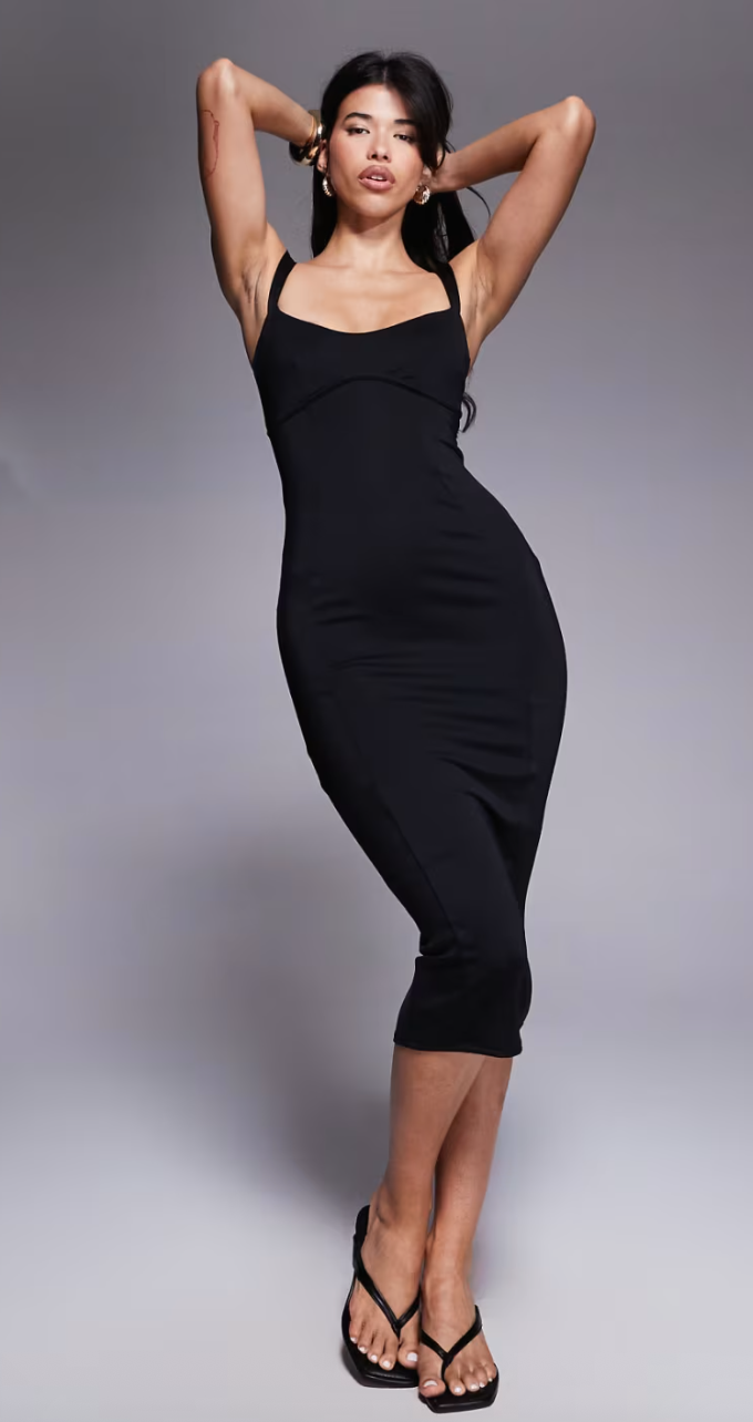 Person in a stylish black sleeveless dress poses confidently with hands behind head, wearing black open-toe sandals