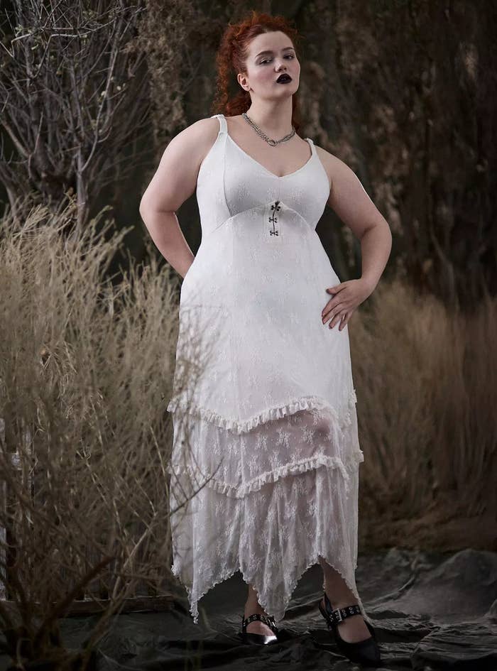 Person in an elegant sleeveless white lace dress with layered asymmetrical hem, posing outdoors among dry foliage