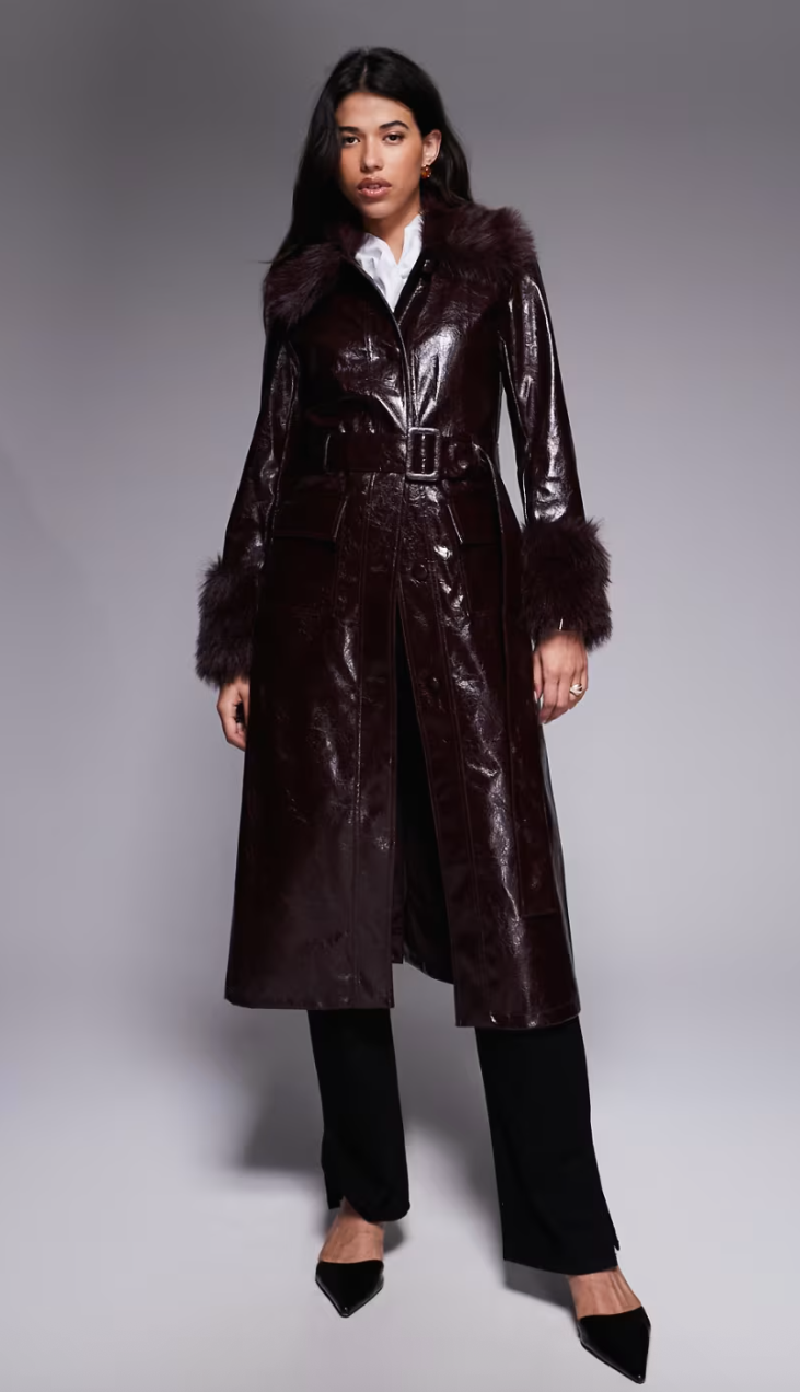 model wearing a sleek, belted faux leather coat with fur trim, paired with black pants and pointed shoes