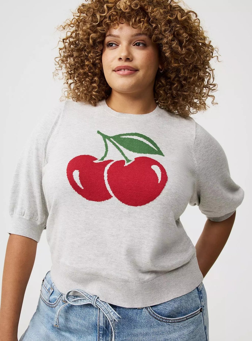 Person wearing a knitted sweater with cherry design, short sleeves, and light-wash jeans, styled for a casual look