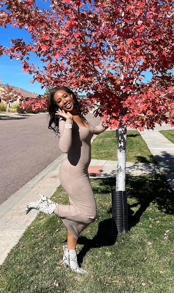 Person poses playfully outdoors by a tree, wearing a form-fitting dress and patterned boots, smiling at the camera