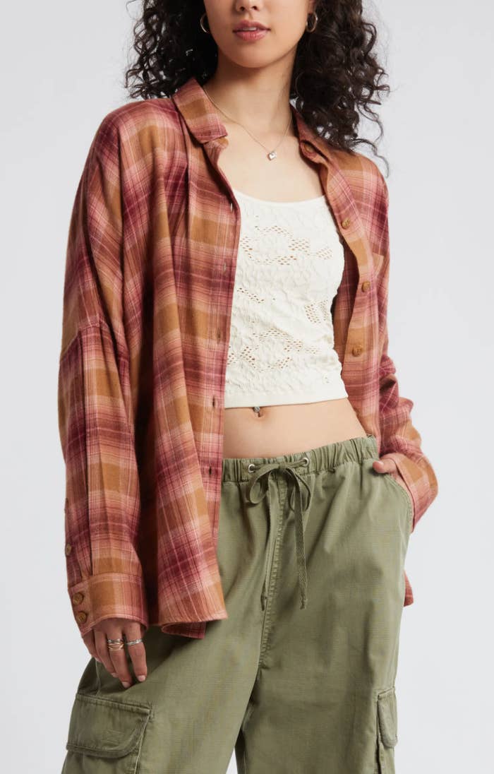 Woman in a plaid shirt over a lace top and green cargo pants is posing with hands in pockets, showing a casual and stylish outfit