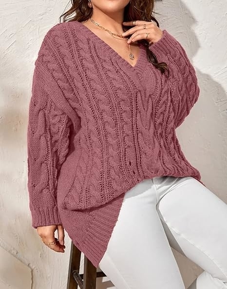 Model wearing a chunky knit V-neck sweater paired with white pants, sitting on a stool.