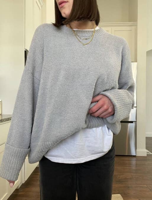 Reviewer posing indoors, wearing the cozy grey oversized sweater with a white shirt underneath and dark pants, accessorized with a layered gold necklace