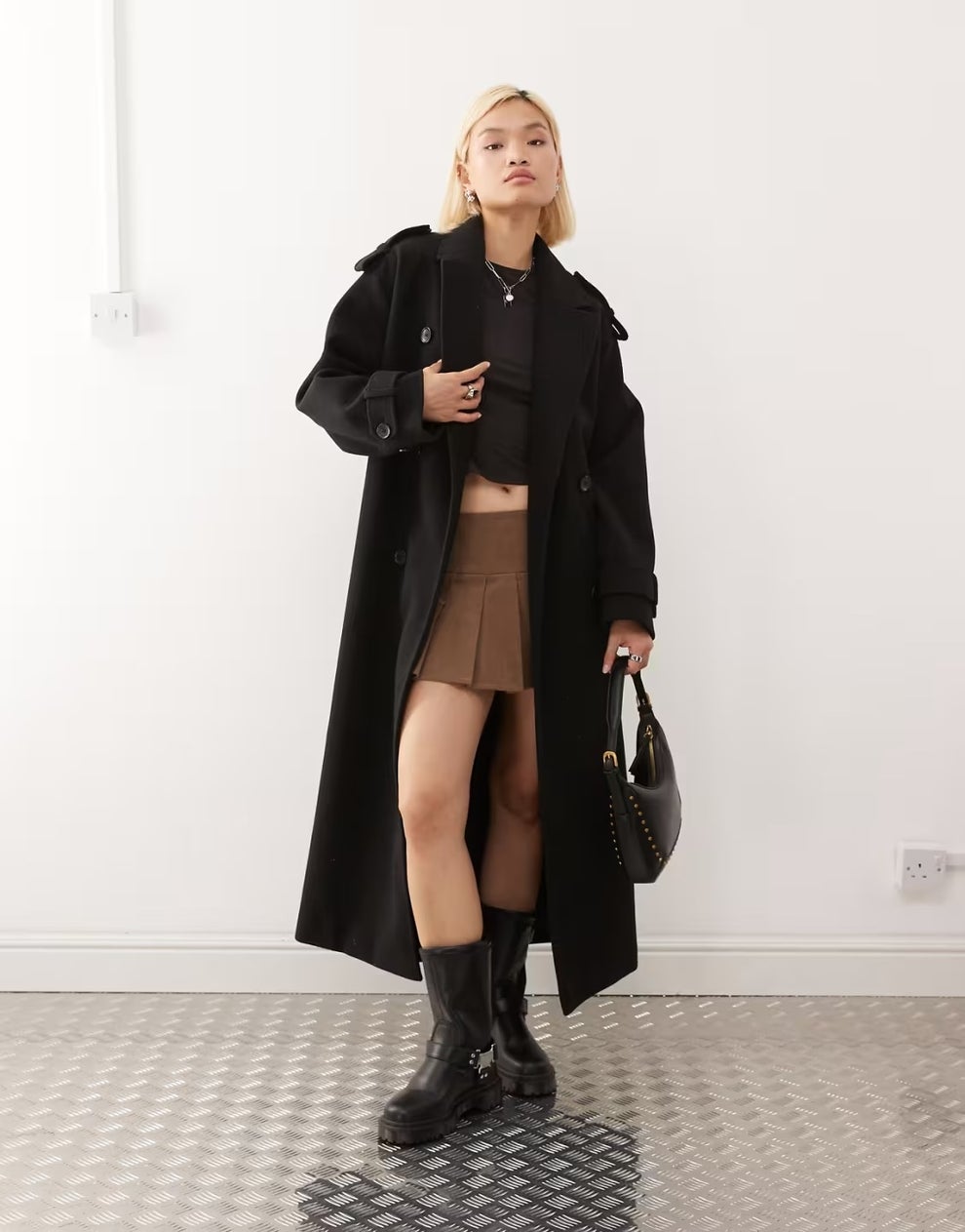 Person in stylish long coat, cropped top, mini skirt, and boots, holding a handbag, standing on a metallic floor