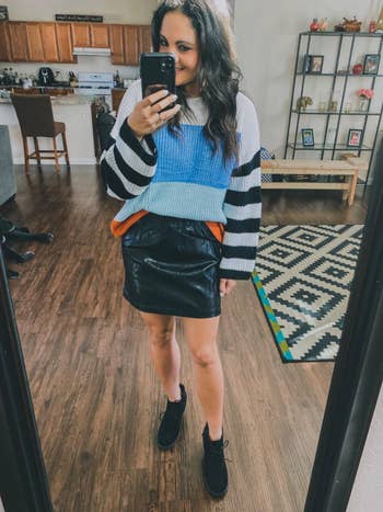 Person in a patterned sweater and leather skirt takes a mirror selfie, showcasing casual fashion in a home setting, relevant to shopping trends